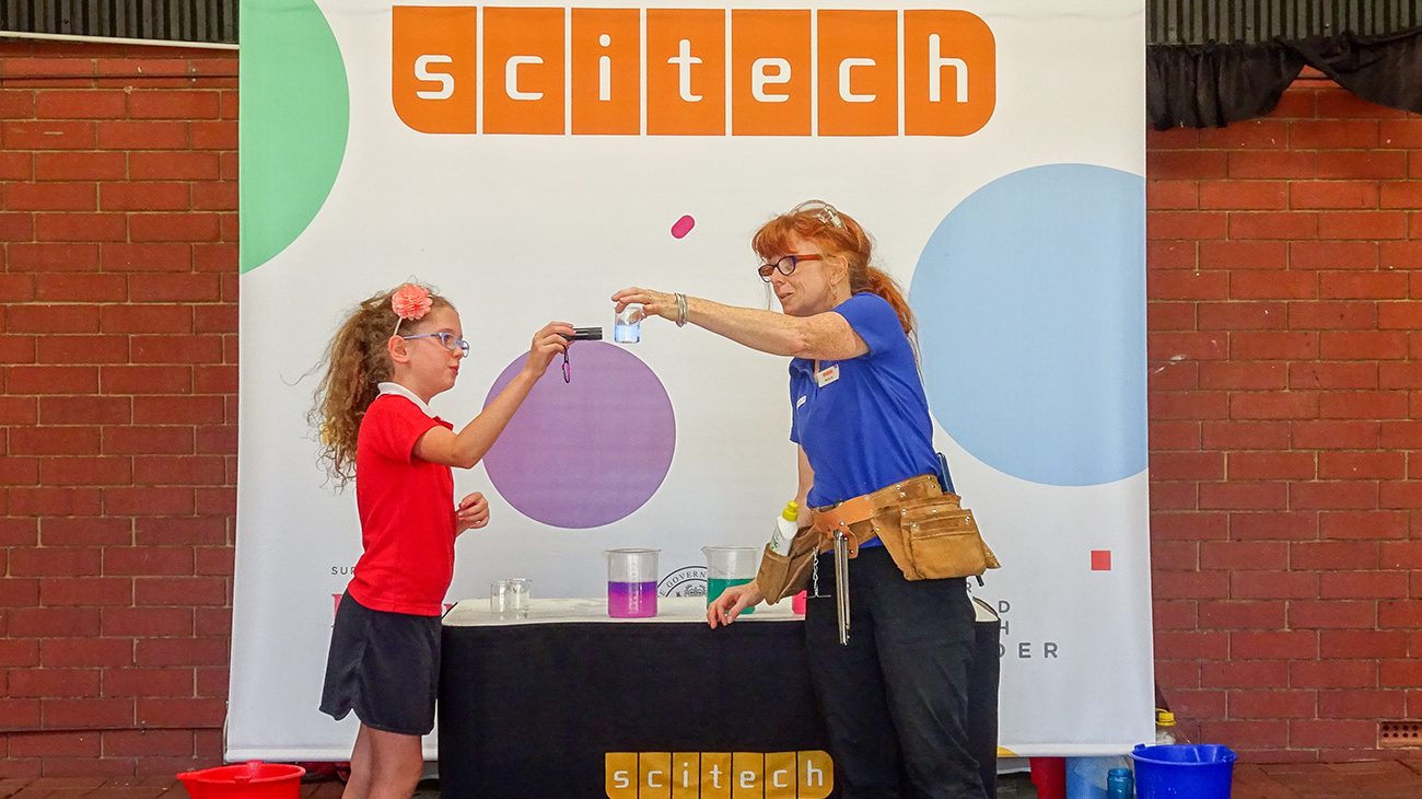 A close up of a Scitech Presenter holding a small beaker of water while a young girl holds an electronic device to it.