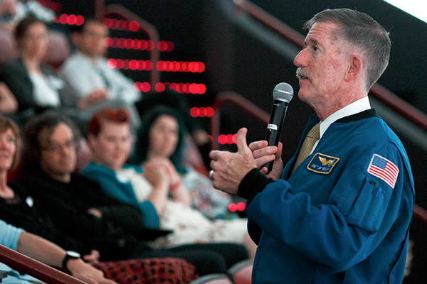 A man wearing an American flag and NASA jacket presenting a speech to a group of people.