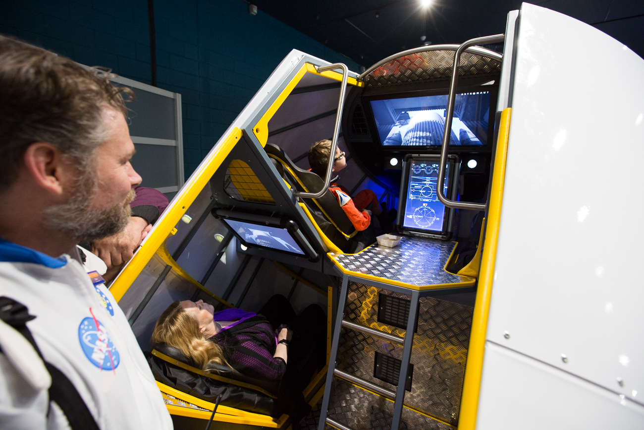 Two people sit inside an exhibit designed to look like a space craft cockpit as a man wearing a NASA jacket looks on