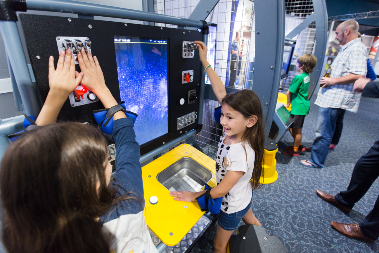 Two young girls playing with the astronaut exhibit at Scitech, pushing switches on a large panel