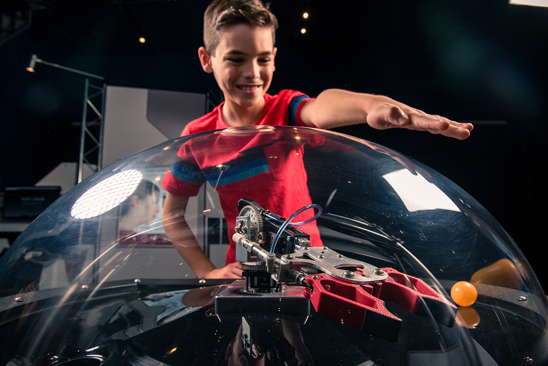 Young boy smiling and waving his hand over a glass dome over a robot and ping pong ball.