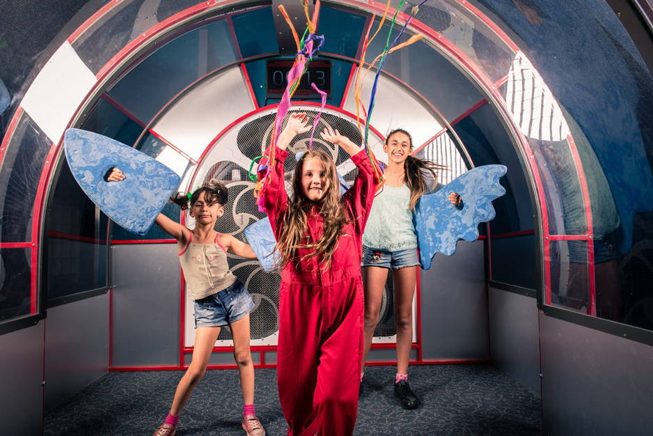 Three teenage girls in a wind tunnel exhibit holding streamers and wings.