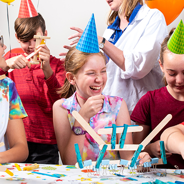 A group of children in birthday hats build objects out of wooden pop sticks and blue pegs