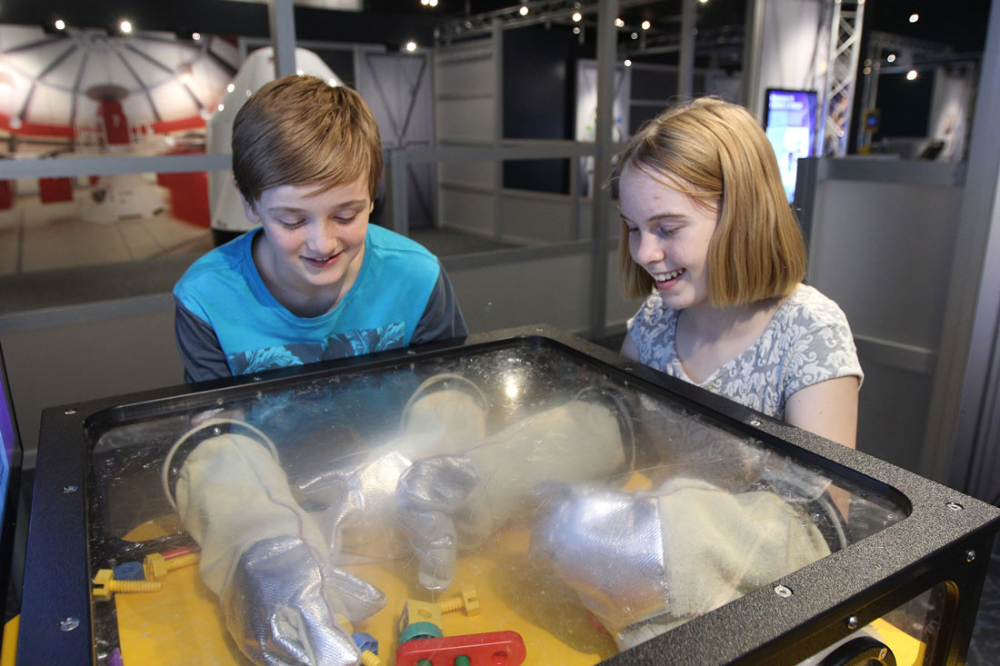 Two children putting their hands in space gloves trying to grab objects at the Astronaut exhibit in Scitech.