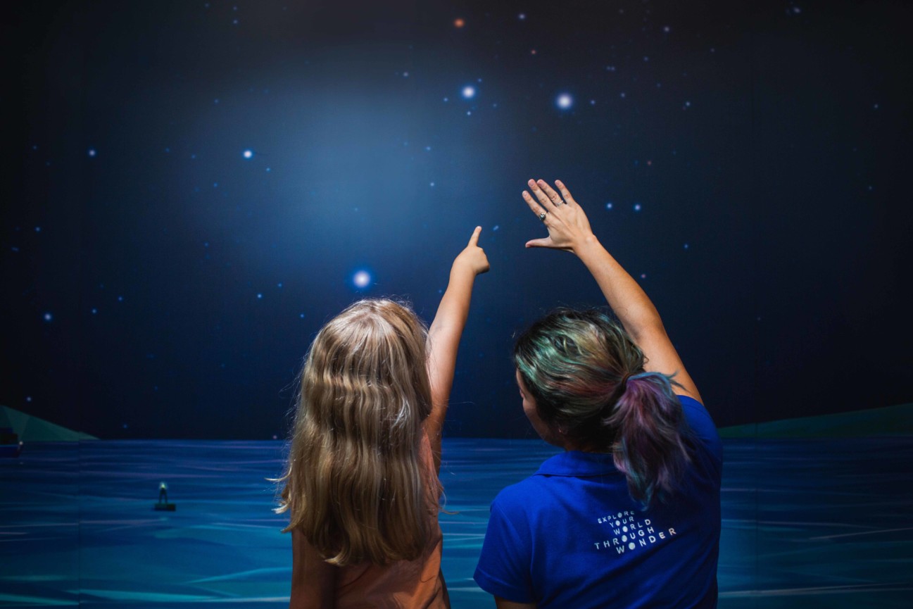 A young girl points to a star on a wall sized image of the night sky over the ocean, with a Scitech staff member guiding them