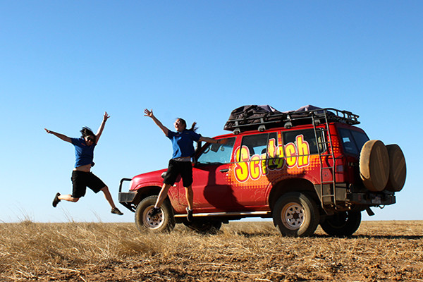 Two scitech outreach staff jump into the air in front of a parked red Scitech branded four wheel drive in regional WA