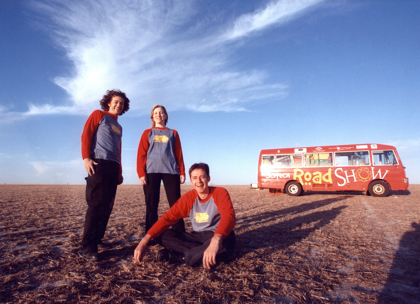 Two scitech staff stand, while a third sits on the ground in a dusty field, with the red 1996 Scitech Road Show Bus in the background The start of Statewide