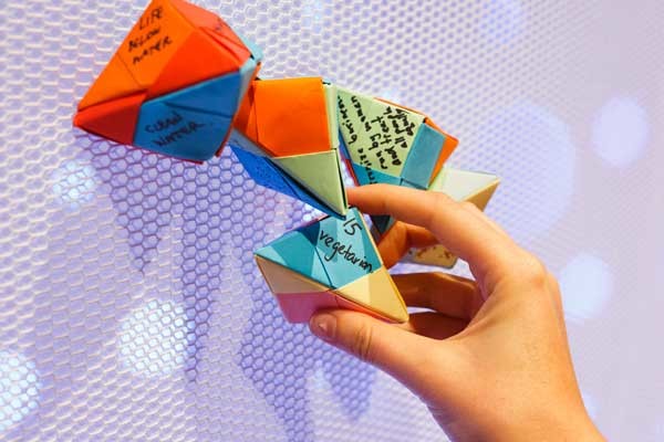 A series of colourful paper origami shapes, folded with writing on each side, attached to a white mesh wall, with a hand grabbing one of the items