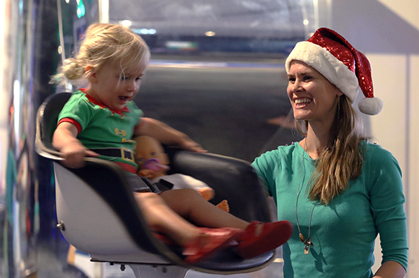 A young girl in an elf outfit with her toy baby sit in a spinning chair while a lady watches her, smiling