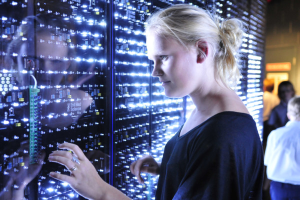 A young woman looking at a wall of light and computer switches.