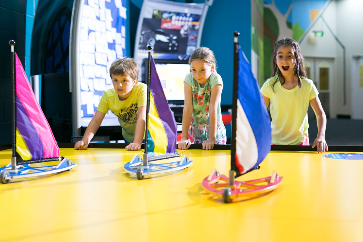 Three children racing land yachts across a table in the Going Places exhibition at Scitech.