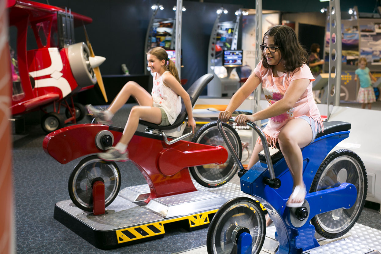 Two teenage girls riding exercise bike models against each other for the Going Places exhibition.