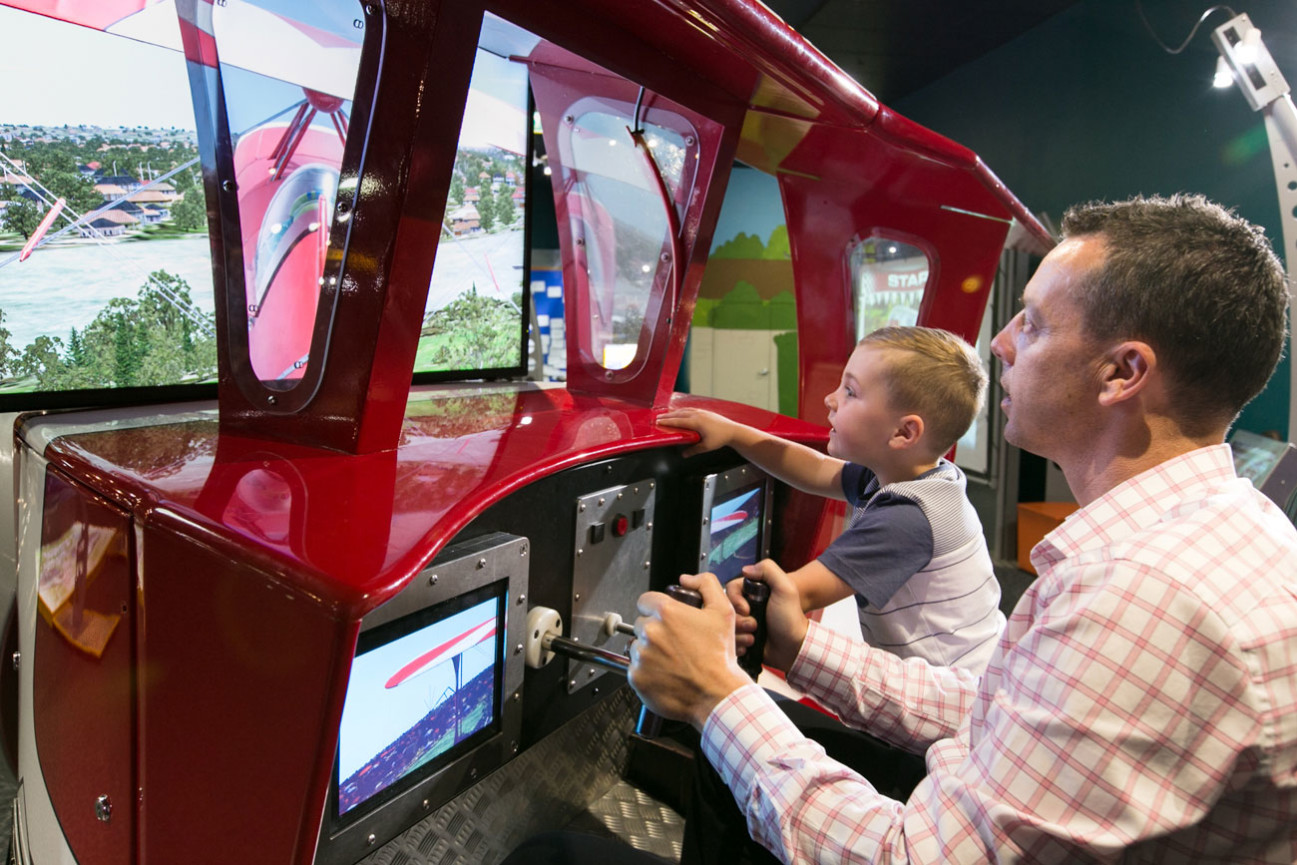 A young boy and father flying a plane in the flight simulator for Going Places Exhibition.