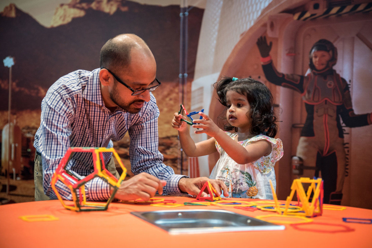 Little girl playing with plastic block toys with her father, a woman in a slim astronaut suit is gesturing behind them.