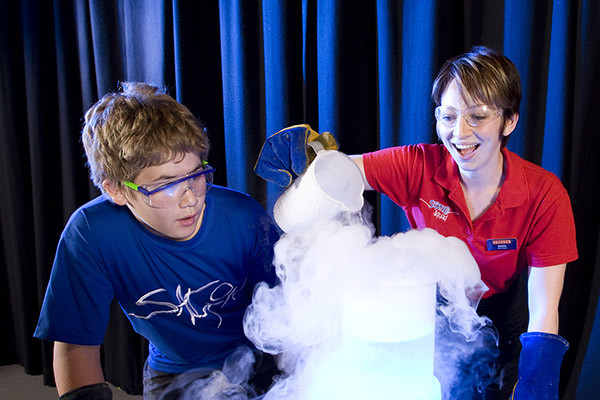 A young woman pouring dry ice into a beaker, a young boy looks on with interest.