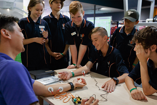 A group of teenagers watching a student and Scitech presenter connect via an electrode circuit
