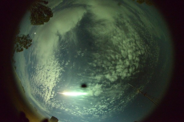 A camera lense effect looking up at a fireball crossing the a cloudy, blue sky.