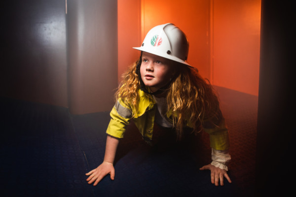 A young girl wearing a firefighters uniform and hard hat crouching on all fours.