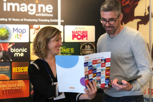 A woman showing a tall man an exhibition brochure at a stall.