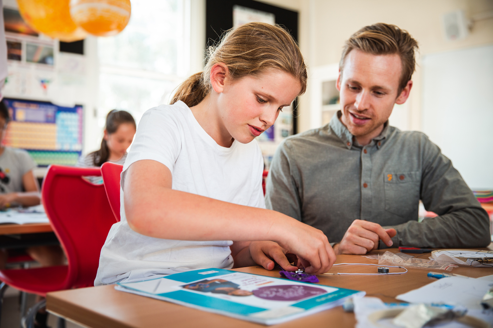 A young adult male supervising a young school girl while she builds a electronic device.