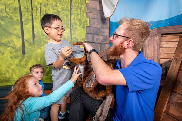 A group of young kids and a science presenter play with a puppet emu.