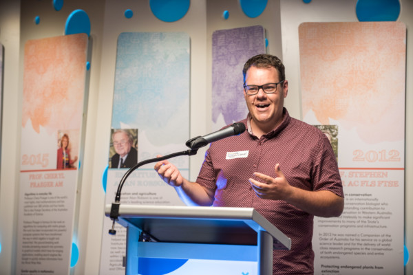 Man talking into microphone, standing in front panels about of WA scientists.