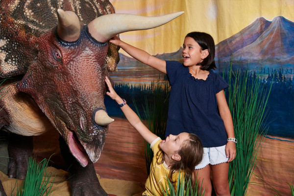 Two young girls patting an animatronic triceratops on the head,