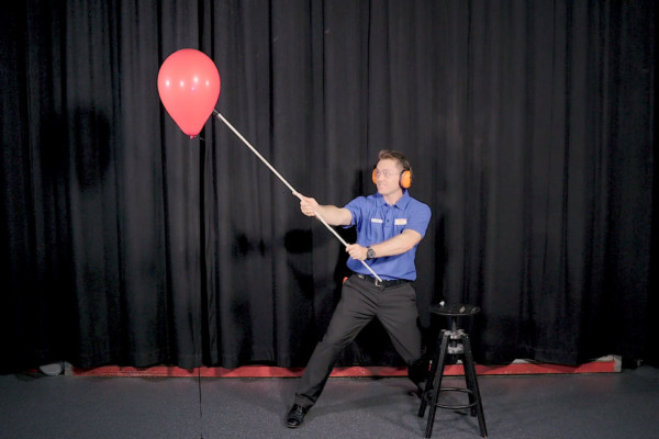 A man about to pop a red balloon with a flame on a stick.