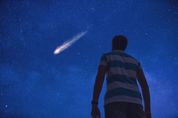 A young boy standing under the night sky looking up at a shooting star.