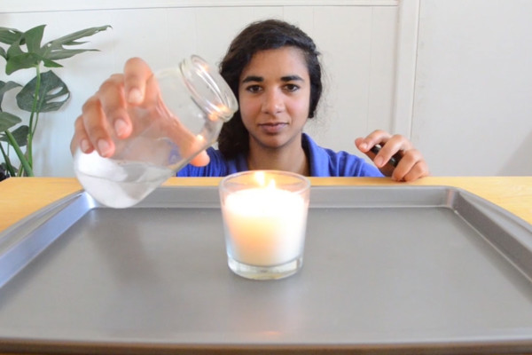A woman in a blue shirt pouring a gas from a jar over a candle's naked flame.