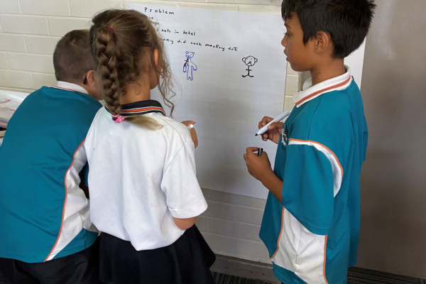 Three primary school children writing on a white board and discussing mathematics.