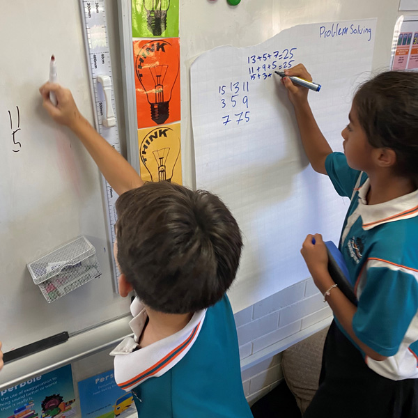 Two primary school children in a classroom writing numbers on a white board.