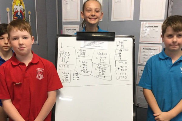 Three primary students standing next to a white board showing math problems they have written.