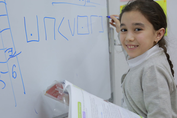 A young girl drawing shapes on a whiteboard whilst holding a textbook and smiling at the camera.