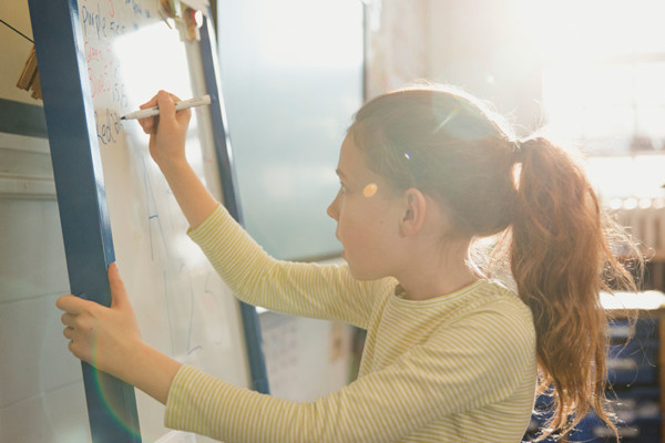 A young girl drawing on a whiteboard with a thoughtful look.