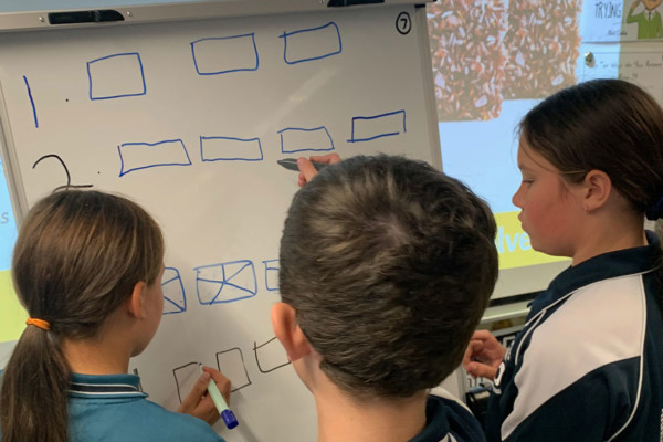 Three primary aged students in a classroom solving math problems on a white board.