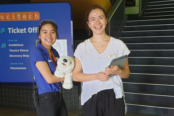 A Scitech presenter holding a stuffed toy with a young female holding an iPad, smiling cheerfully.