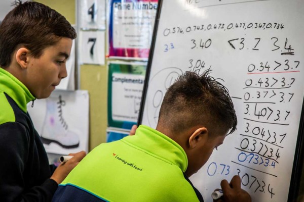 Two primary student boys writing math symbols on a small whiteboard.