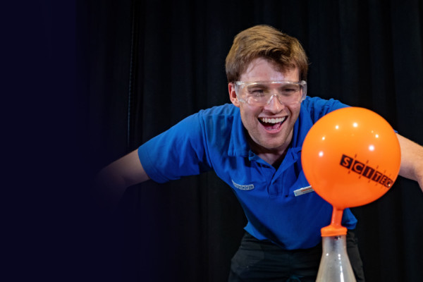 A Scitech presenter wearing a blue short laughs as an orange balloon, which has been placed over the top of a conical flask, fills up