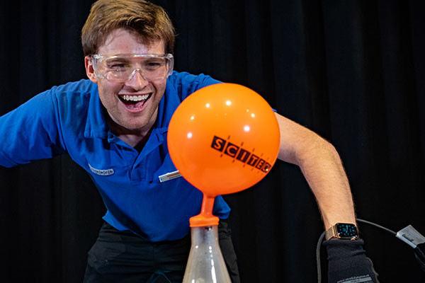 A Scitech presenter wearing a blue short laughs as an orange balloon, which has been placed over the top of a conical flask, fills up