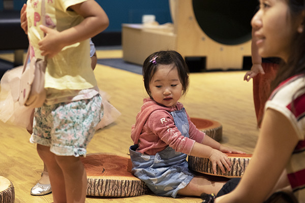 A toddler wearing a pink hoodie sits on a cushion that looks like a piece of log wood