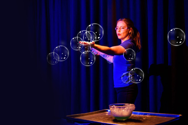 A female presenter wearing a blue Scitech t-shirt holds her hand out in front of her. There are bubbles on her hand and around her, and a bowl of soapy detergent on a table in front of her