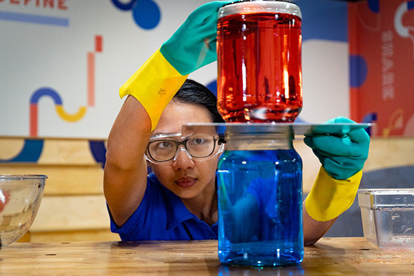 A science presenter looks closely at two jars stacked on top of each other, one containing blue water and one containing red water, as part of an experiment into water density