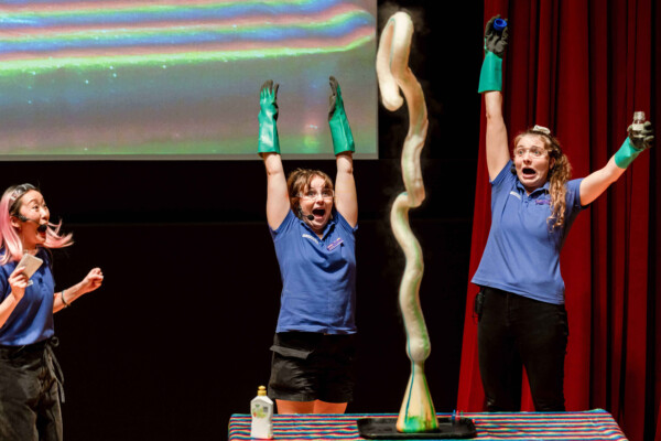 Three people cheer as a chemical reaction causes brown and green foam to shoot upwards from a beaker