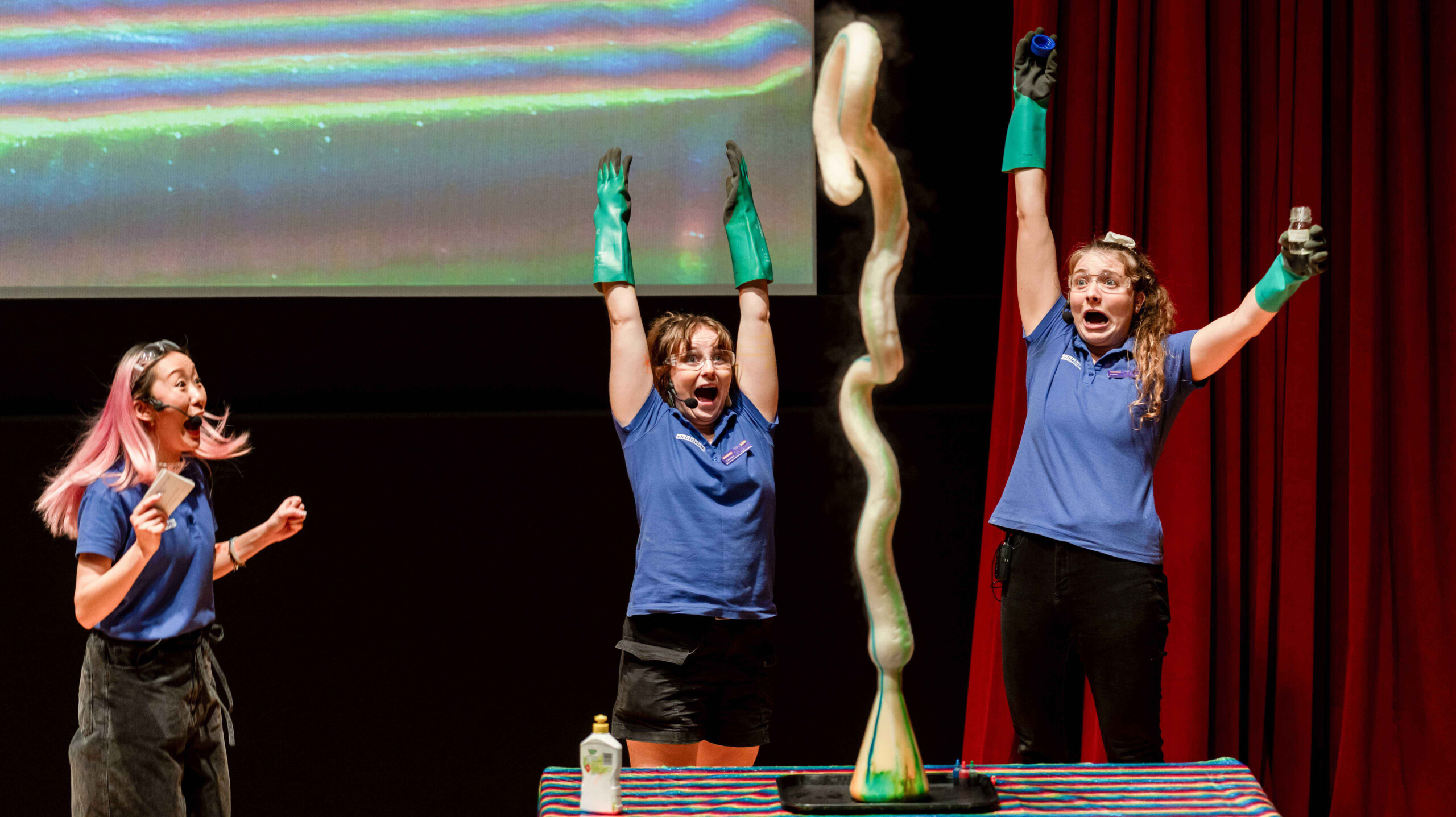 Three people cheer as a chemical reaction causes brown and green foam to shoot upwards from a beaker