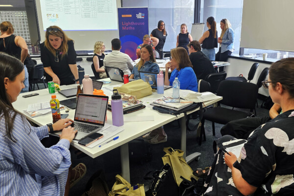 Primary school teachers gather around a desk with their notebooks and laptop as Lighthouse Maths coach Gesla explains an activity.